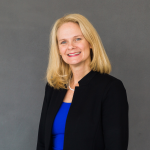 A woman with blonde hair in a blue shirt and black blazer smiles at the camera in front of a grey backdrop.