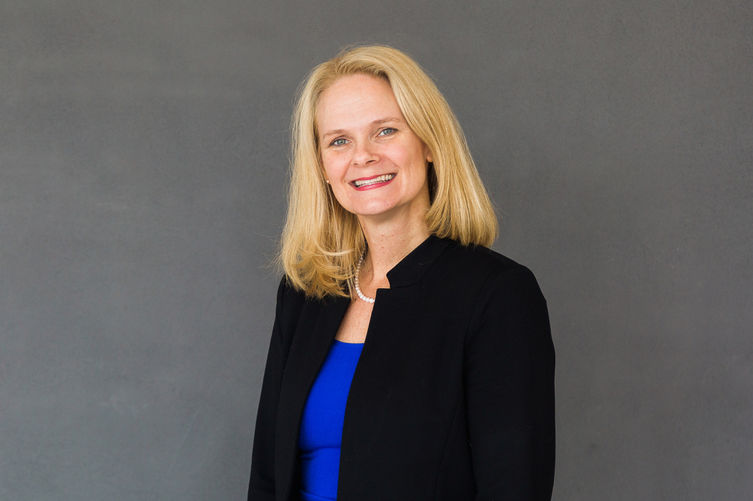 A woman with blonde hair in a blue shirt and black blazer smiles at the camera in front of a grey backdrop.