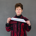A woman with short hair in a red ad black patterned shirt holds a sign that reads "#Museums Are Not Neutral.