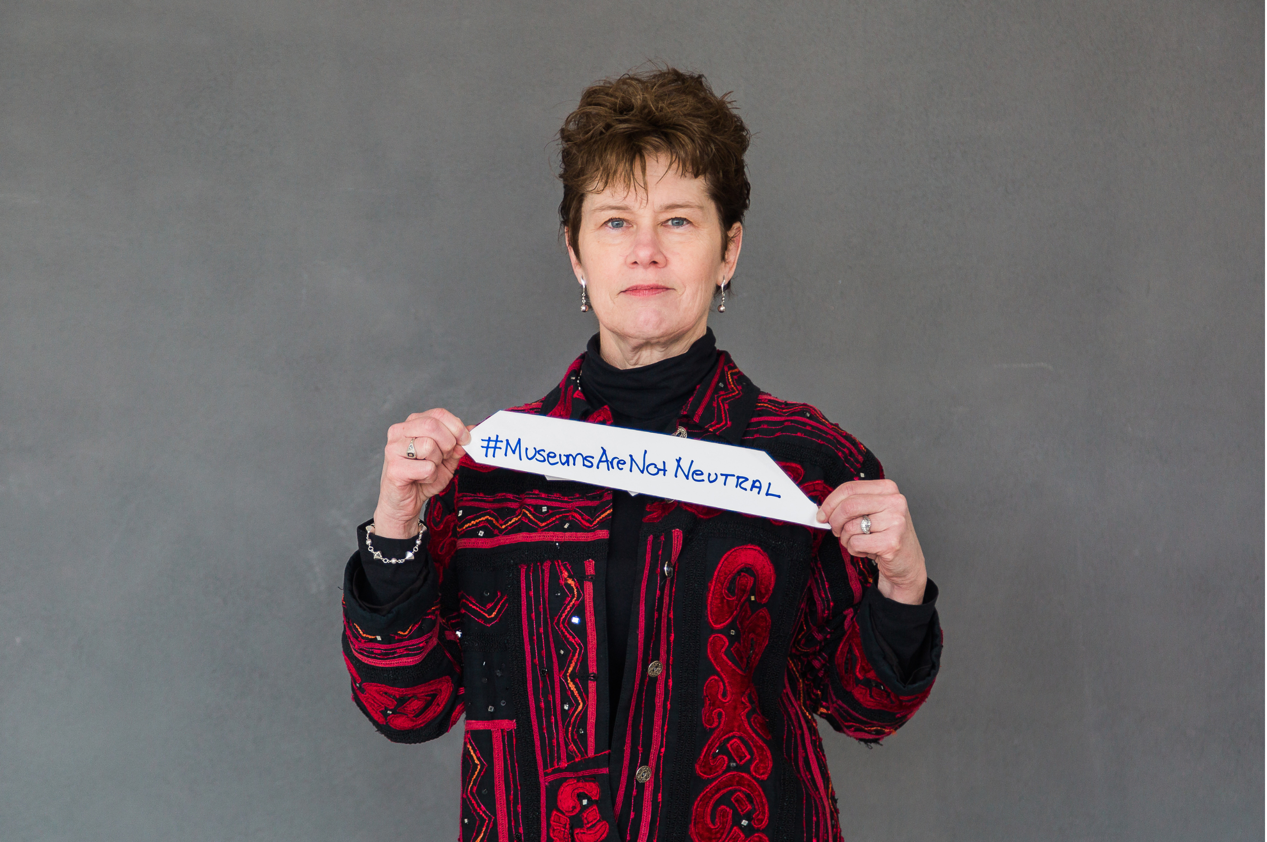 A woman with short hair in a red ad black patterned shirt holds a sign that reads "#Museums Are Not Neutral.