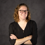 A woman with short wavy hair in glasses and a black three-quarter sleeve shirt smiles at the camera.