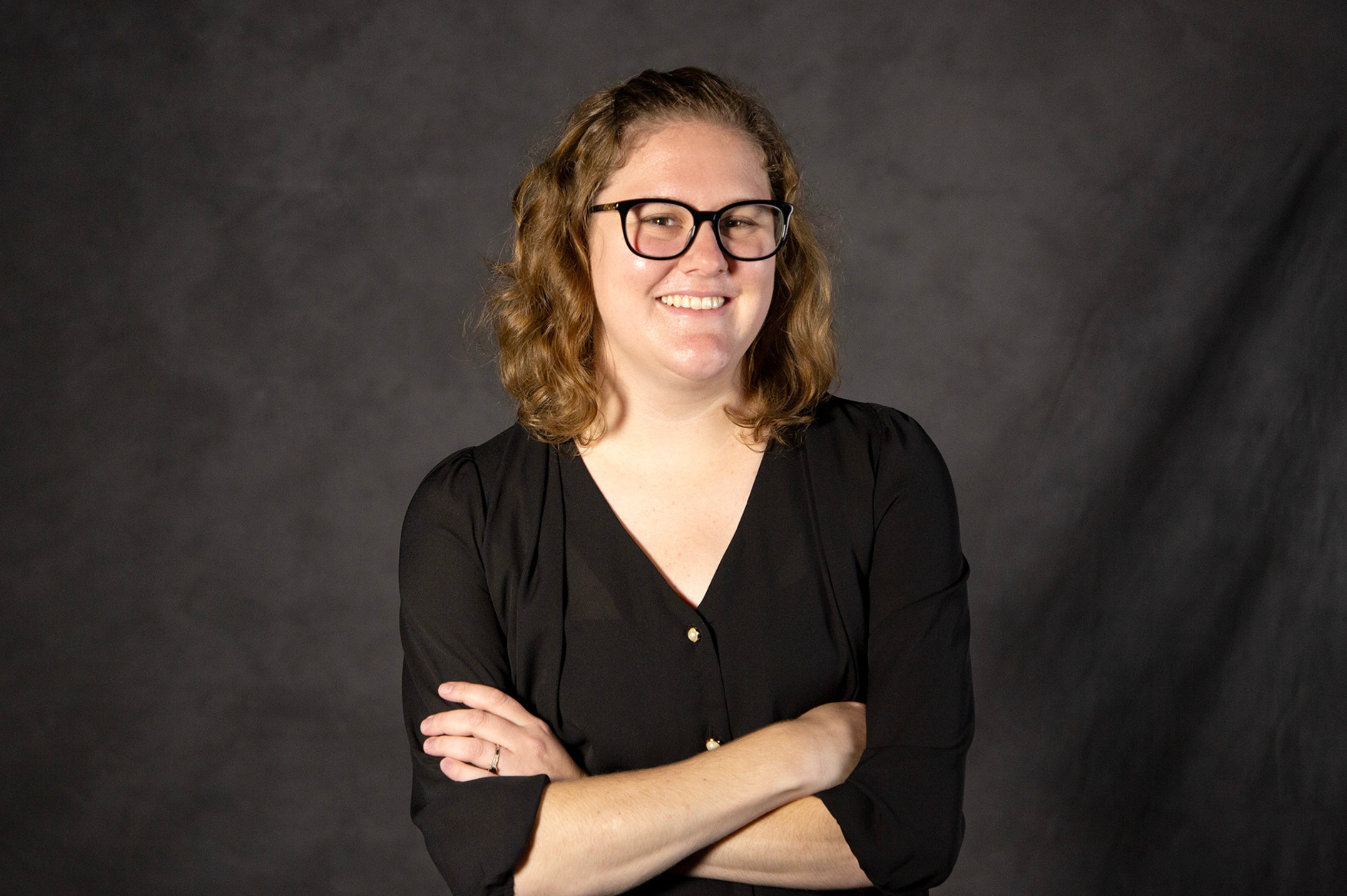 A woman with short wavy hair in glasses and a black three-quarter sleeve shirt smiles at the camera.