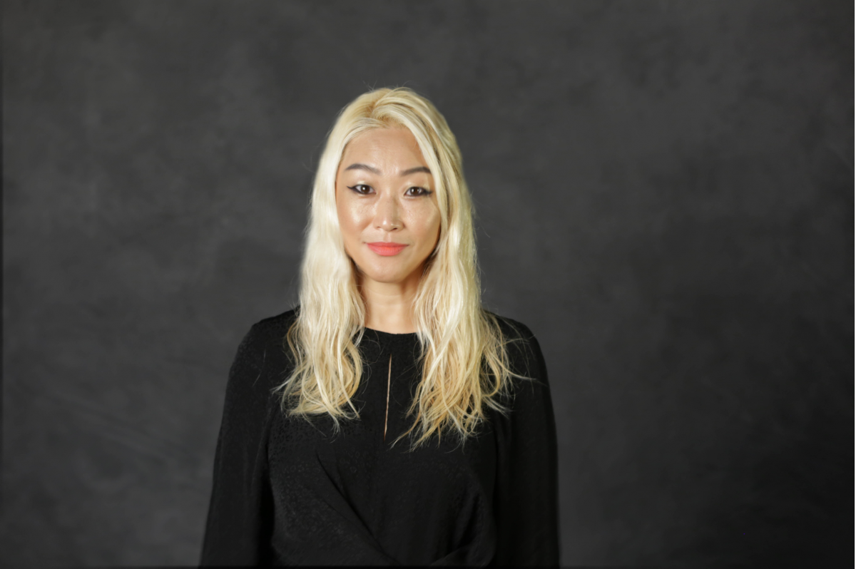 A woman with long blonde hair in a long sleeve shirt smiles at the camera in front of a dark grey back drop.
