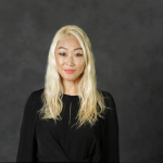 A woman with long blonde hair in a long sleeve shirt smiles at the camera in front of a dark grey back drop.