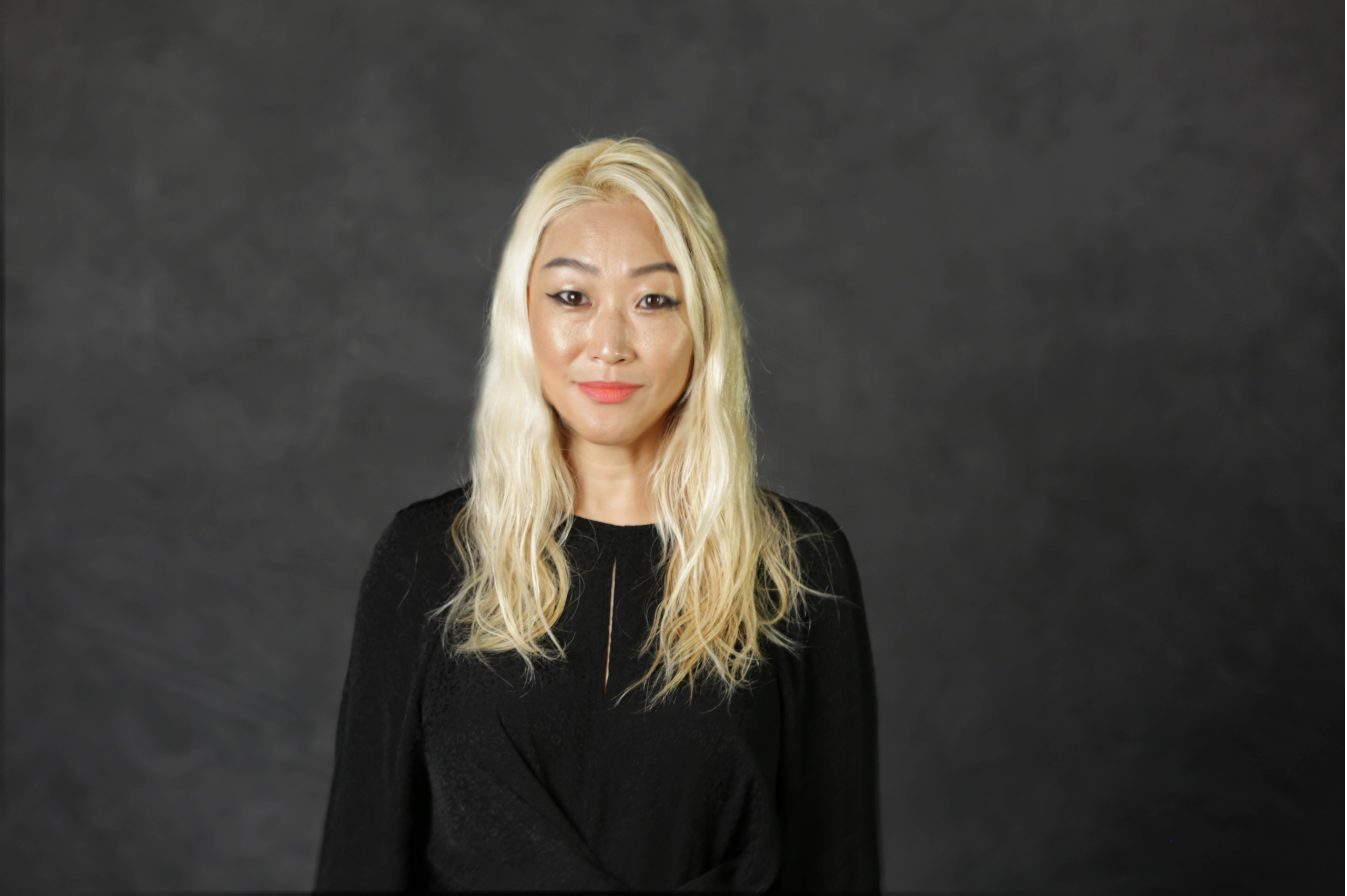 A woman with long blonde hair in a long sleeve shirt smiles at the camera in front of a dark grey back drop.