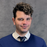 A man with brown curly hair in a blue sweater, white collared shirt, and tie smiles at the camera in front of a grey back drop.