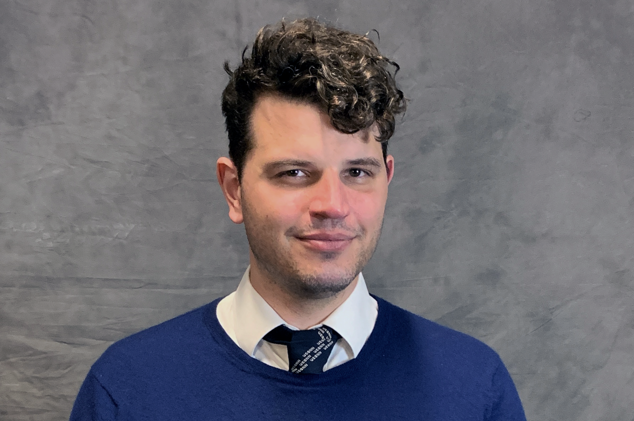 A man with brown curly hair in a blue sweater, white collared shirt, and tie smiles at the camera in front of a grey back drop.