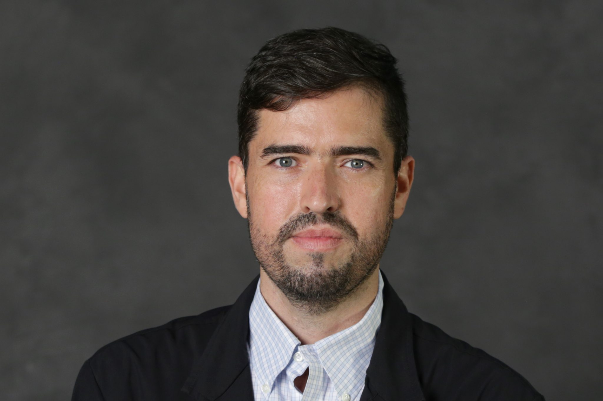A man with dark brown hair in a black suit jacket and light blue, plaid button down looks at the camera in front of a gray back drop.