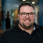 A man with brunette hair and facial hair wearing dark, tortoise shell glasses and a black polo smiles at the camera.