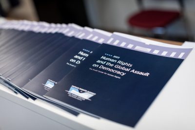 A stack of dark navy booklets read "Human Rights and the Global Assault on Democracy."