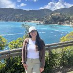 A woman with long, dark hair wearing tan pants, a white shirt, light brown cardigan and baseball hat smiles at the camera in front of a tropical background of blue water, green bushes and rolling hills. 