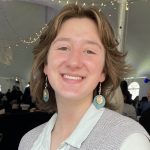 A woman with short, blonde hair wearing a light lavender vest, white collared shirt and dangling earrings smiles at the camera. 