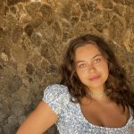 A woman with long, wavy, brunette hair wearing a white and blue patterned short sleeve top looks at the camera. 
