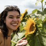 A woman with dark, shoulder length hair turns to smile at the camera next to a large sunflower. 