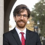 A man with brunette hair and facial hair wearing a black suit, white button down, and red polka dot tie leans against a wall while smiling at the camera.