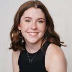 A woman with shoulder length brunette hair wearing a black tank top leans slightly toward the camera and smiles. 