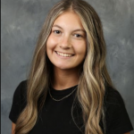 A woman with long blonde hair wearing a black shirt and small chain necklace smiles at the camera in front of a grey backdrop. 