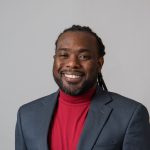 A man wearing a dark grey suit jacket and red turtle neck smiles at the camera in front of a light grey backdrop.