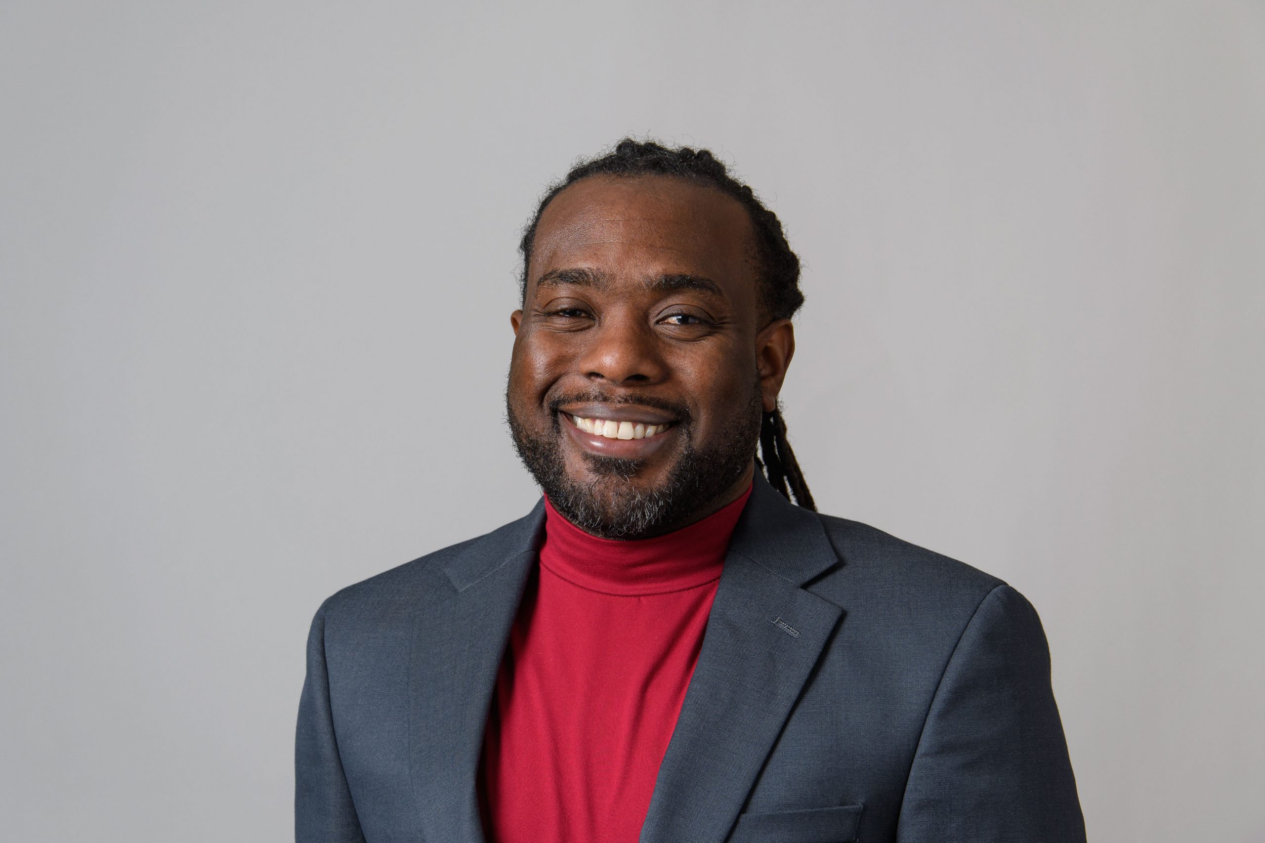 A man wearing a dark grey suit jacket and red turtle neck smiles at the camera in front of a light grey backdrop.