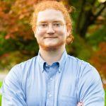A man with ginger hair pulled back has facial hair and is wearing a light blue button down. He smiles at the camera in front of a tree whose leaves are changing colors for fall.