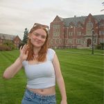 A woman with ginger hair wearing a white shirt and jeans smiles at the camera in front of a brick building on a green lawn.
