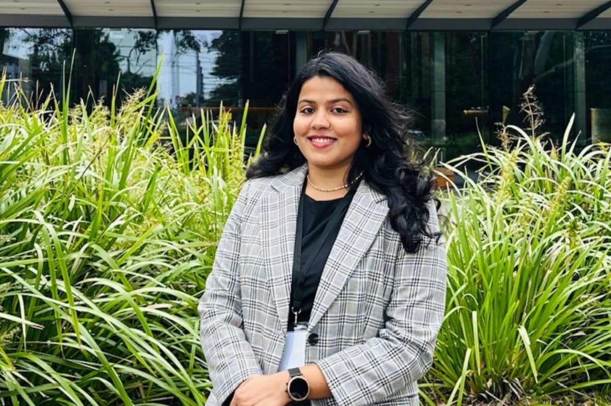 A woman with long, black hair wearing a light grey, plaid blazer and black under shirt smiles at the camera in front or tall, green bushes.