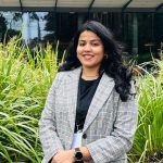 A woman with long, black hair wearing a light grey, plaid blazer and black under shirt smiles at the camera in front or tall, green bushes.