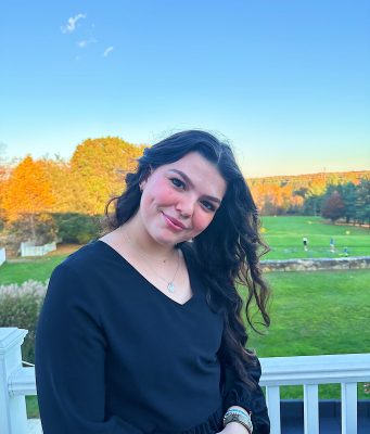 A woman with long, dark brunette hair wearing a black, long sleeve shirt leans her head to the left while smiling at the camera in front of a green lawn and trees.