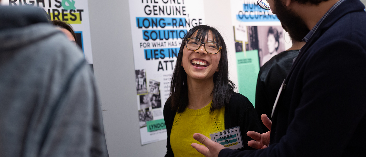 A woman in a yellow shirt and glasses is laughing while socializing at a poster session.