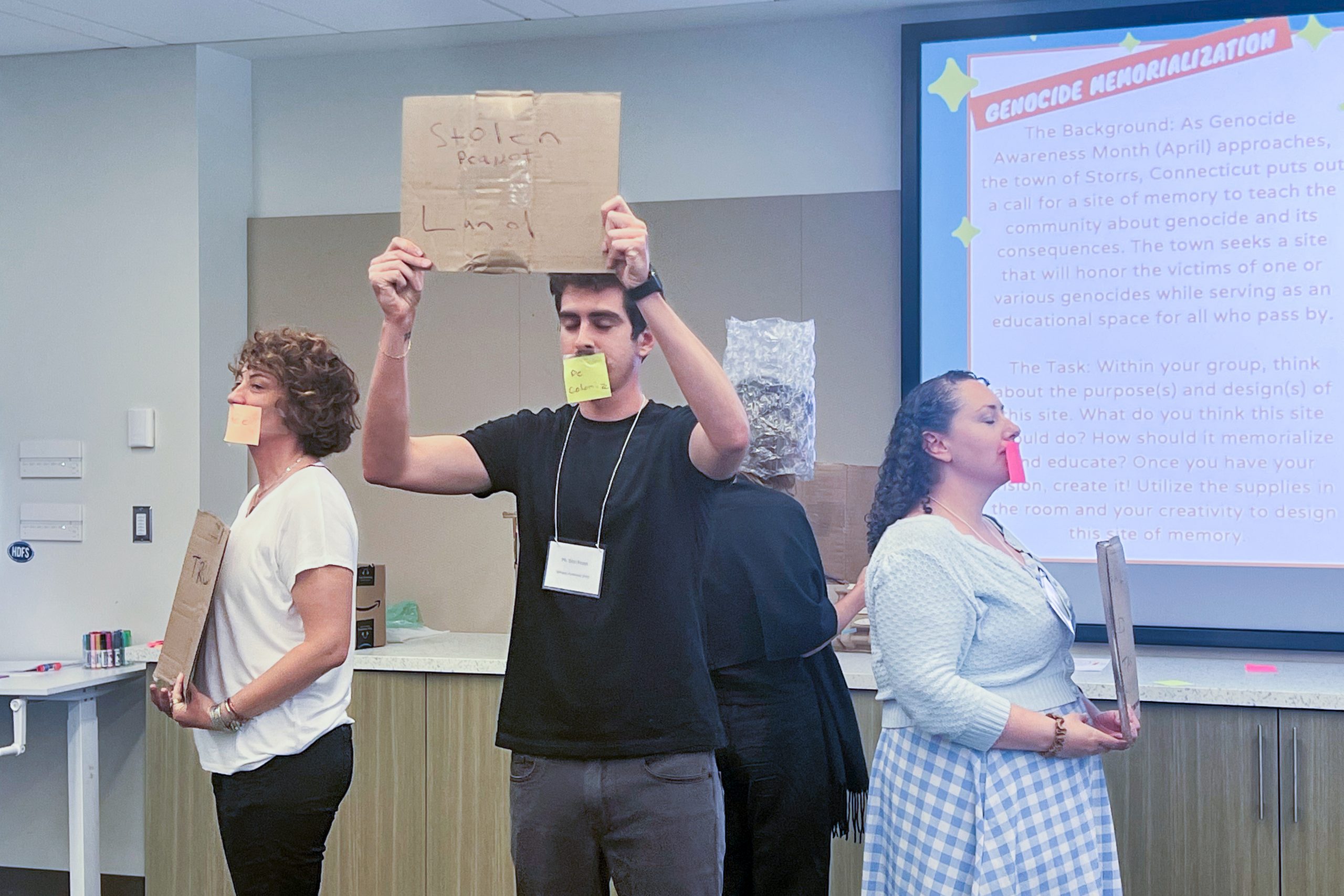 Participants of the Summer Seminar for Genocide Studies and Prevention stand holding signs with paper over their mouths to present memorials they created during a session